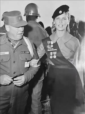 A smiling white woman in a military costume walks arm in arm with a white man in an army uniform